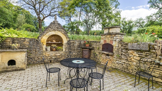 Un patio de piedra con una mesa redonda y sillas, rodeado de muros de piedra y vegetación.