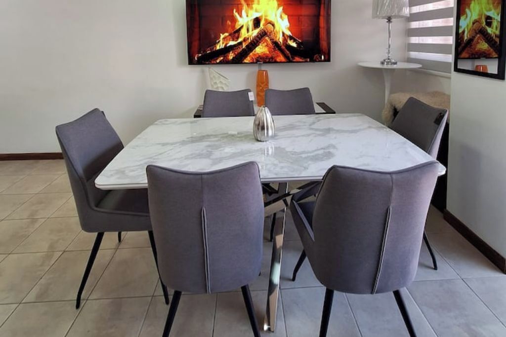 A dining area with a marble table, grey upholstered chairs, a vase centerpiece, and a wall-mounted flat-screen TV displaying a fireplace.