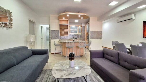 A modern living room with a grey sofa, a marble coffee table, and a kitchen area with a bar and seating. - Luxury & Location, Beautiful Apartment in Los2Caminos-Boleita (Caracas)