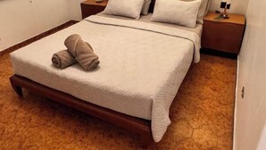 Una cama bien hecha con una toalla doblada, un cabecero de madera y una mesita de noche con lámpara y teléfono.