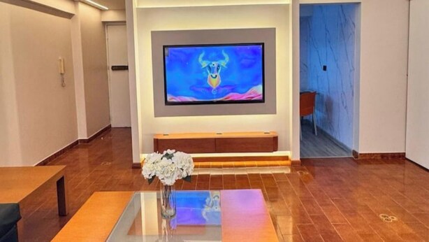 A modern living room with a flat-screen TV mounted on the wall, a wooden coffee table with a vase of flowers, and a blue-tiled floor.