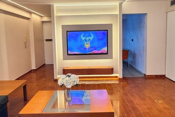 A modern living room with a flat-screen TV mounted on the wall, a wooden coffee table with a vase of flowers, and a blue-tiled floor.
