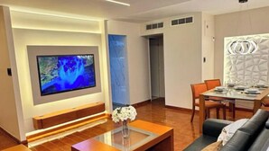 Una sala de estar moderna con un televisor de pantalla plana montado en la pared, una mesa de centro de vidrio con un jarrón de flores, un sofá de cuero negro y una zona de comedor con mesa y sillas.