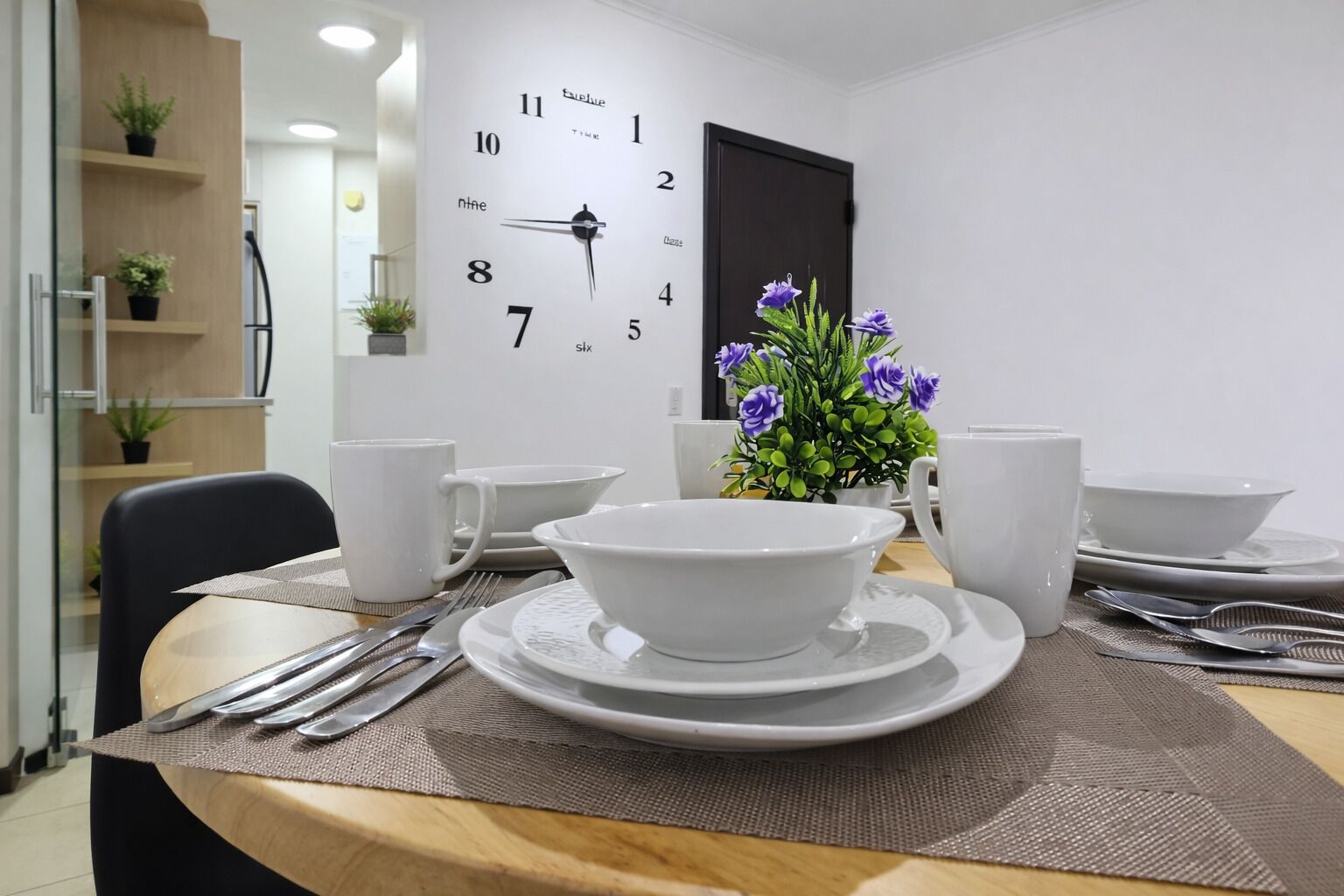 A dining area with a table set for four, including bowls, cups, and cutlery. A clock is mounted on the wall, and there are potted plants on shelves.