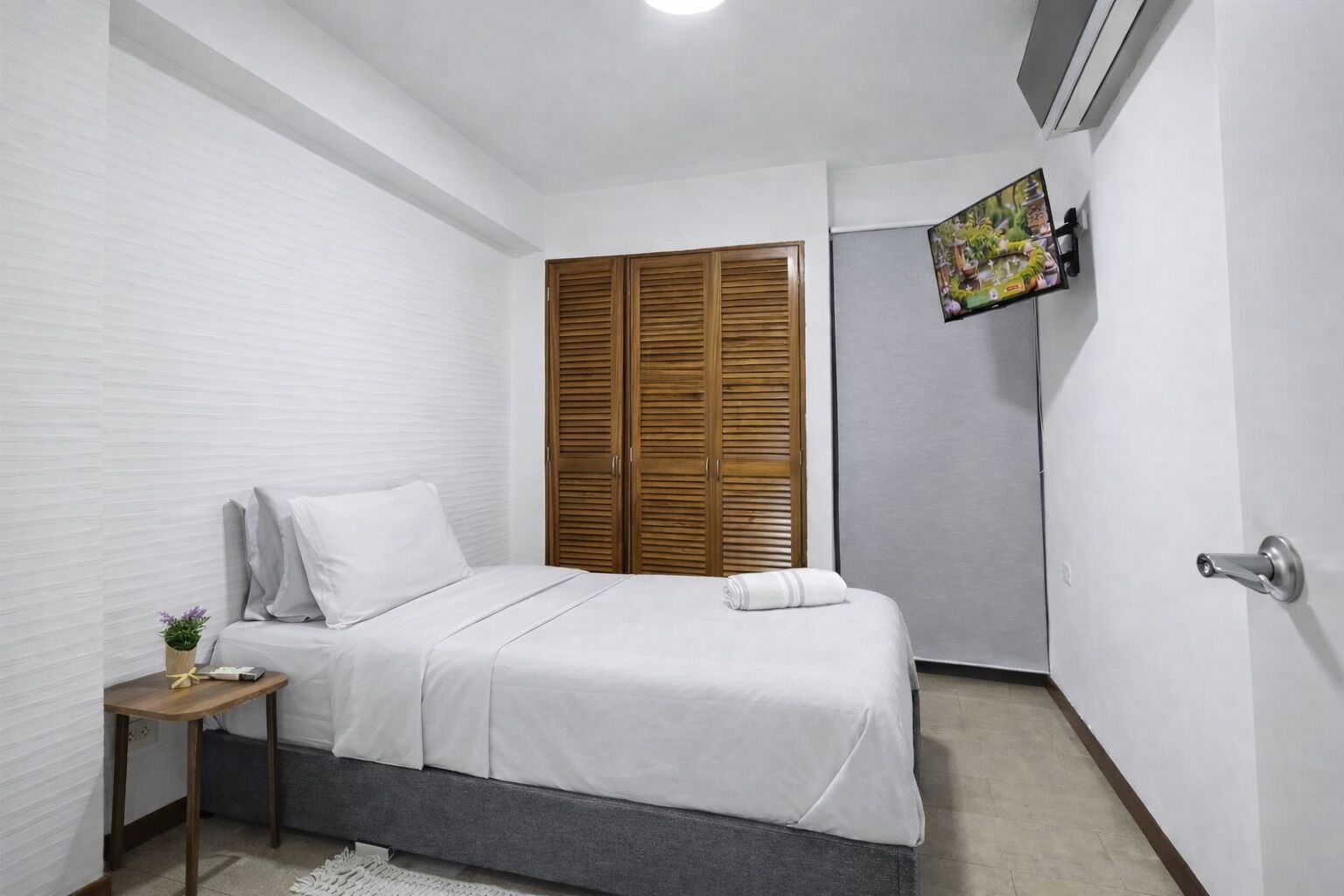 A bedroom with a bed, a nightstand, a wooden wardrobe, and a wall-mounted TV.