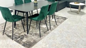 Ein Essbereich mit einem rechteckigen Tisch, sechs bequemen grünen Stühlen, einem gemusterten Teppich und einem grauen Sofa im Hintergrund.