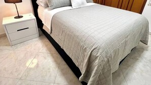 Ein ordentlich bezogenes Bett mit einer gesteppten Tagesdecke, ein Nachttisch mit Lampe und eine Kommode.
