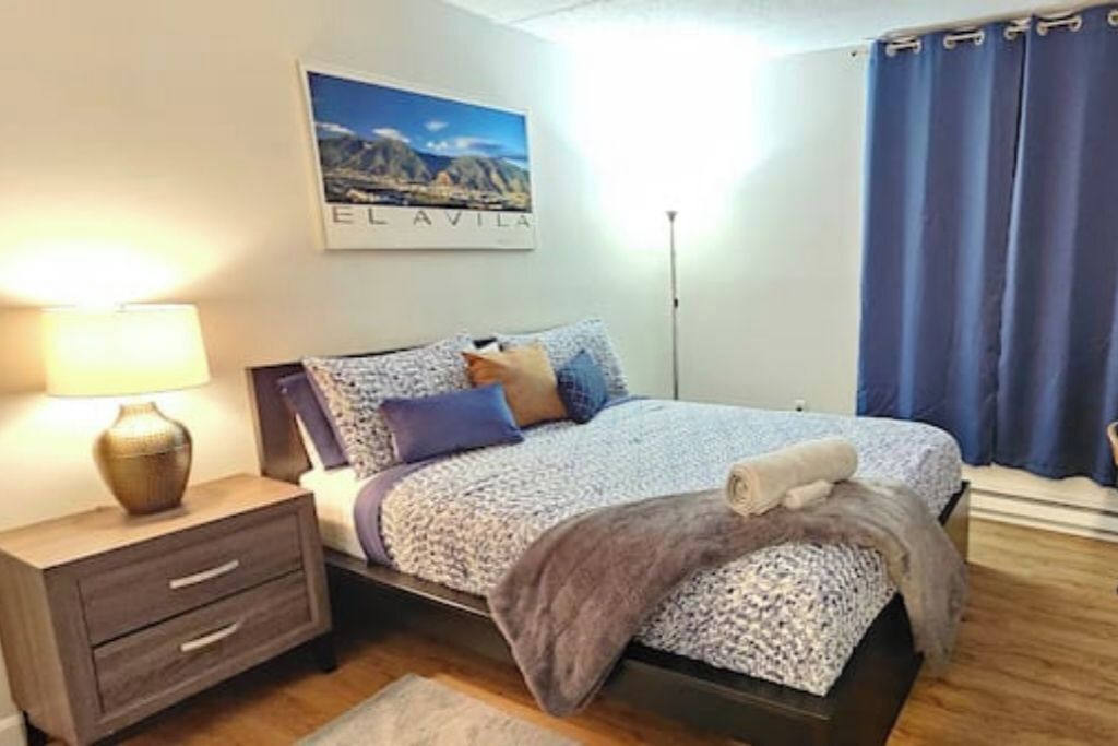 Une chambre bien rangée, avec un lit, une table de chevet sur laquelle se trouve une lampe, un tableau encadré au mur et une fenêtre avec des rideaux.