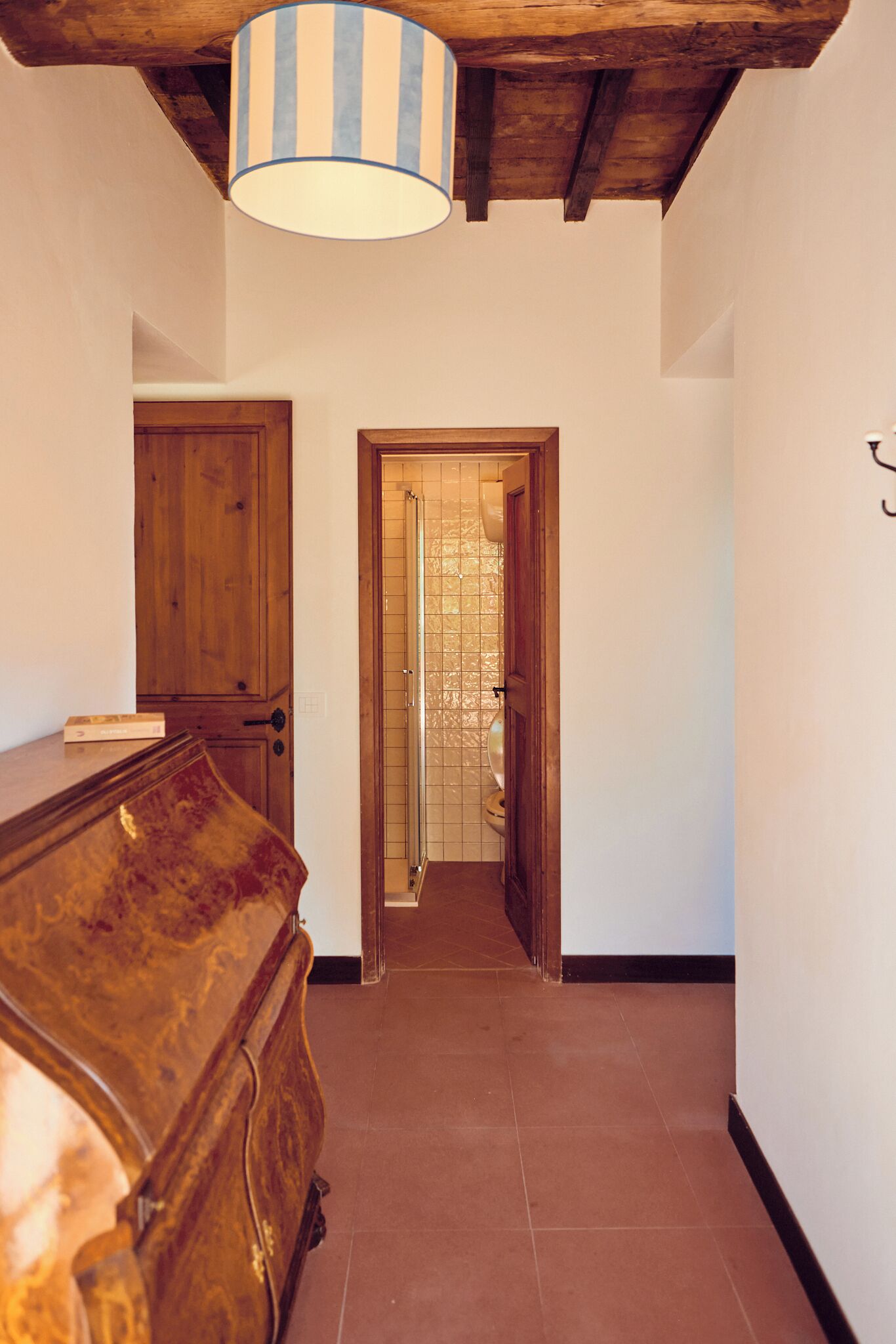 Un couloir avec une commode en bois, une lampe à rayures et un sol carrelé, qui mène à une porte dotée d’un panneau de verre.