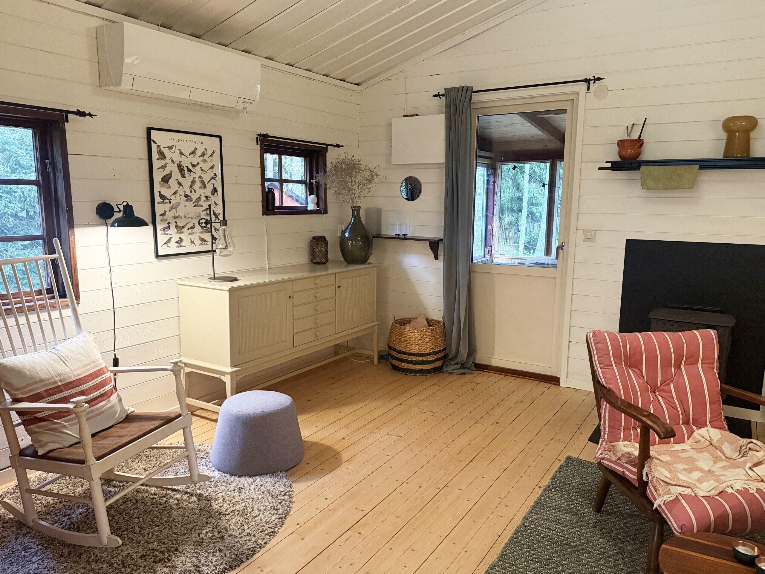 A cozy room with a wooden floor, a rocking chair, a striped armchair, a small ottoman, a white cabinet, a framed picture, a vase, a basket, and a fireplace.