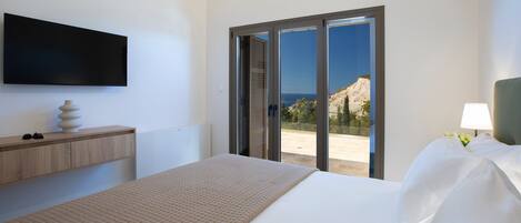 A modern hotel room with a bed, a flat-screen TV, a wooden bedside table, a lamp, and a view of the outdoors.