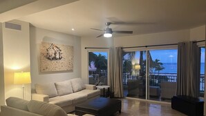 Un salon moderne avec un canapé beige, une table basse et un pouf noir. On y trouve un ventilateur de plafond, un tableau accroché au mur et une vue sur l’océan grâce à de grandes portes coulissantes.