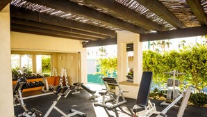 Une salle de sport couverte équipée de divers appareils d’exercice, notamment des haltères et des machines de musculation, et offrant une vue sur la verdure environnante.