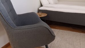 Una habitación moderna con un sillón gris, una cama con ropa de cama blanca y un cuadro abstracto enmarcado en la pared.