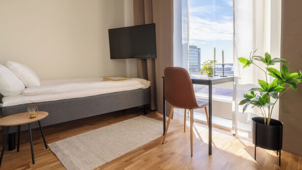 Una habitación de hotel moderna con una cama, un televisor montado en la pared, una mesita con un vaso, una silla y una planta en maceta.