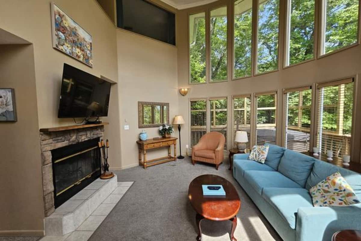 A spacious living room with a fireplace, a television, a sofa, and large windows offering a view of trees.