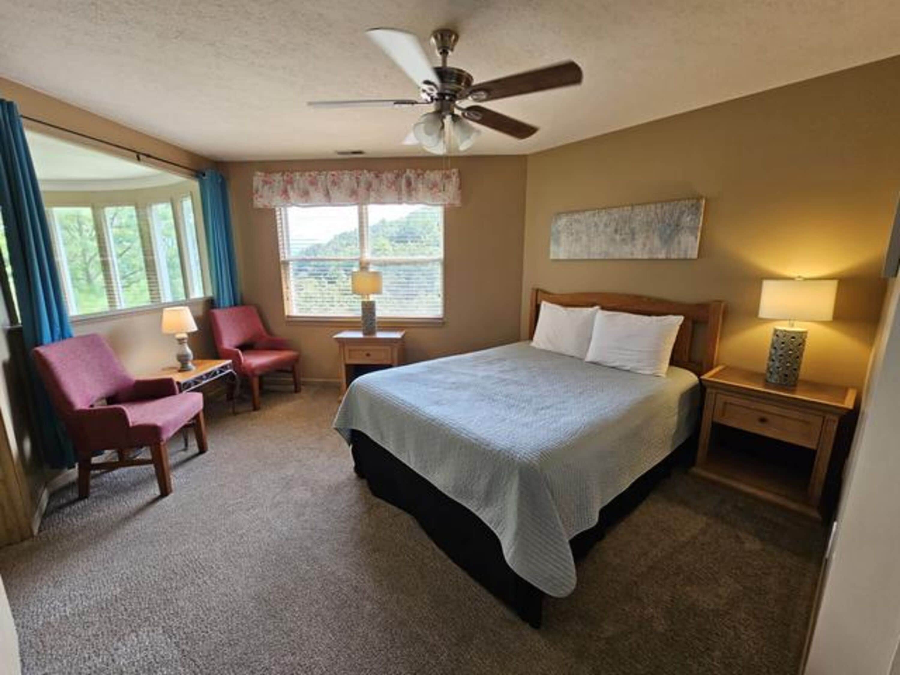 A hotel room with a bed, a chair, a desk, a lamp, a ceiling fan, and a window with curtains.