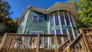 Une maison à deux étages, avec une façade incurvée, une terrasse en bois et un ciel bleu clair en arrière-plan.
