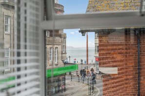 Blick durch ein Fenster auf eine Stadtstraße mit Gebäuden, einer Fußgängerampel und einem entfernten Gewässer.