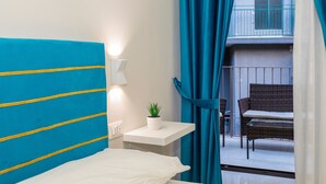 A hotel room with a bed, a small table with a plant, a window with curtains, and a balcony with a chair.