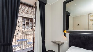 A hotel room with a bed, a small table, a mirror, and a view of an outdoor area with a brick wall and a balcony with chairs.