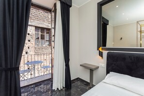 A hotel room with a bed, a small table, a mirror, and a view of an outdoor area with a brick wall and a balcony with chairs.