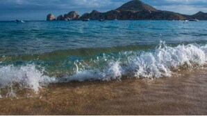 A beach with clear water, waves, and rocky formations in the distance. - Las Mañanitas LM E3302 (San José del Cabo)