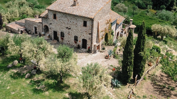 Un bâtiment en pierre, surmonté d’un toit de tuiles, entouré d’une végétation luxuriante et avec des collines en arrière-plan.