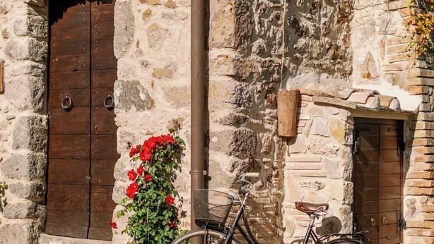 A stone building with a wooden door, a bicycle, and a flower pot. - West Way (Tolfa)