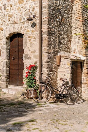 A stone building with a wooden door, a bicycle, and a flower pot. - West Way (Tolfa)