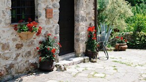 A stone building with two arched windows and a door, surrounded by potted plants and a cobblestone path. - West Way (Tolfa)