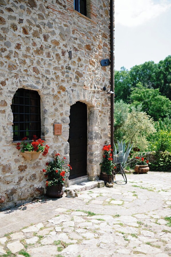 A stone building with two arched windows and a door, surrounded by potted plants and a cobblestone path.