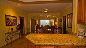 A kitchen with a granite countertop, wooden cabinets, a dining area with chairs, and a view of the ocean through large windows. - Las Mañanitas LM E3303 (San José del Cabo)