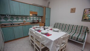 A compact kitchen with teal cabinets, a dining table set for four, and a patterned sofa in the corner. - Cozy Apartment Near the Beach (Rosolina)