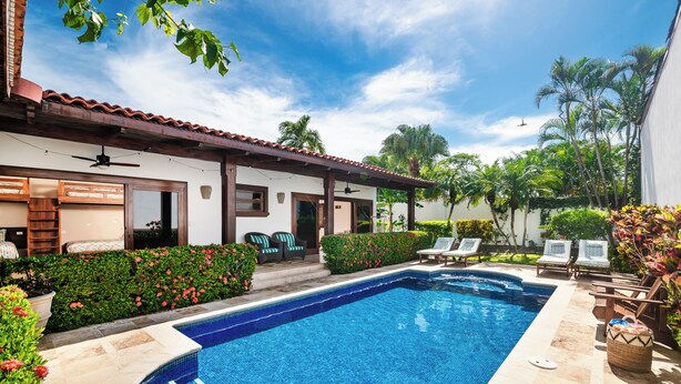 A swimming pool with a tiled deck, surrounded by lounge chairs and a small building with a tiled roof.