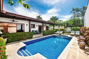 A swimming pool with a tiled deck, surrounded by lounge chairs and a small building with a tiled roof.