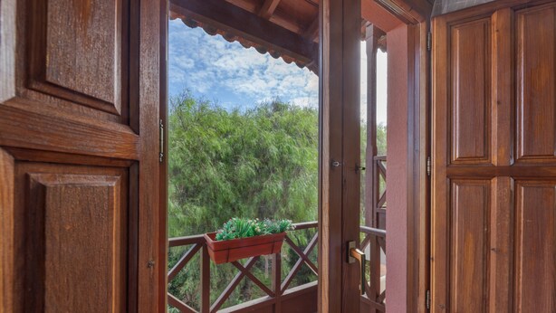 Eine offene Holztür, die auf einen Balkon mit Blick auf Grün und einen klaren Himmel führt.