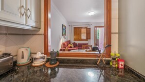 Eine Küche mit Toaster, Wasserkocher und Kaffeemaschine auf der Arbeitsfläche, einem Holzkorpus und Blick in ein Wohnzimmer mit Sofa und Gemälde.