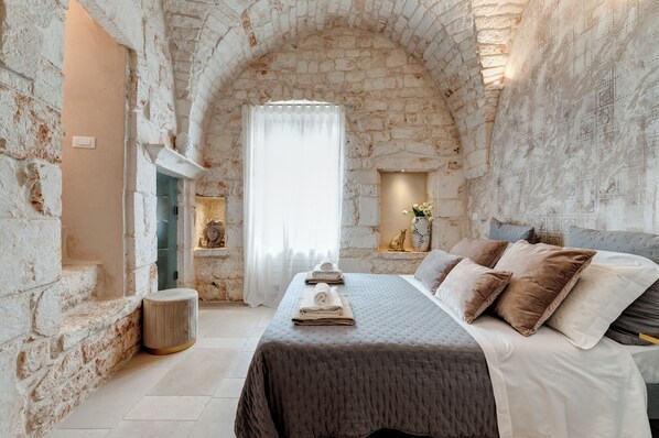 A stone-walled room with a bed, a small stool, and a window with curtains.