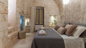 A room with stone walls, a bed with pillows, a window with curtains, and a small statue on a shelf.