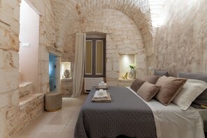 A room with a stone wall, a bed with a grey bedspread, a small stool, and a window with curtains.