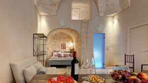 A cozy room with a bed, a dining table with food, a wine bottle, and a fruit bowl.