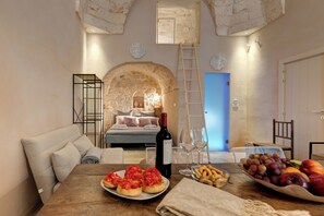 A cozy room with a bed, a dining table with food, a wine bottle, and a fruit bowl.