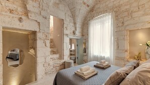 A room with exposed brick walls, a bed with neatly folded towels, a small stool, and a window with sheer curtains.