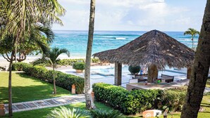 A pool area with a thatched roof structure, surrounded by palm trees and a view of the ocean.