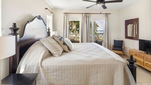 A bedroom with a large bed, a ceiling fan, a wooden dresser, a TV, and a balcony with a view of the ocean.