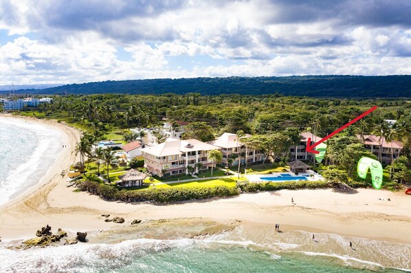 Um hotel na praia com piscina, vários edifícios e vista para o oceano e as colinas.