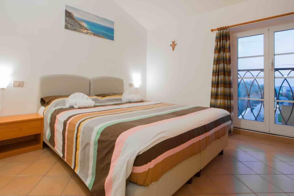 Ein Hotelzimmer mit einem Bett, einem Nachttisch, einem Gemälde, einem Kruzifix an der Wand und einem Fenster mit Vorhängen.