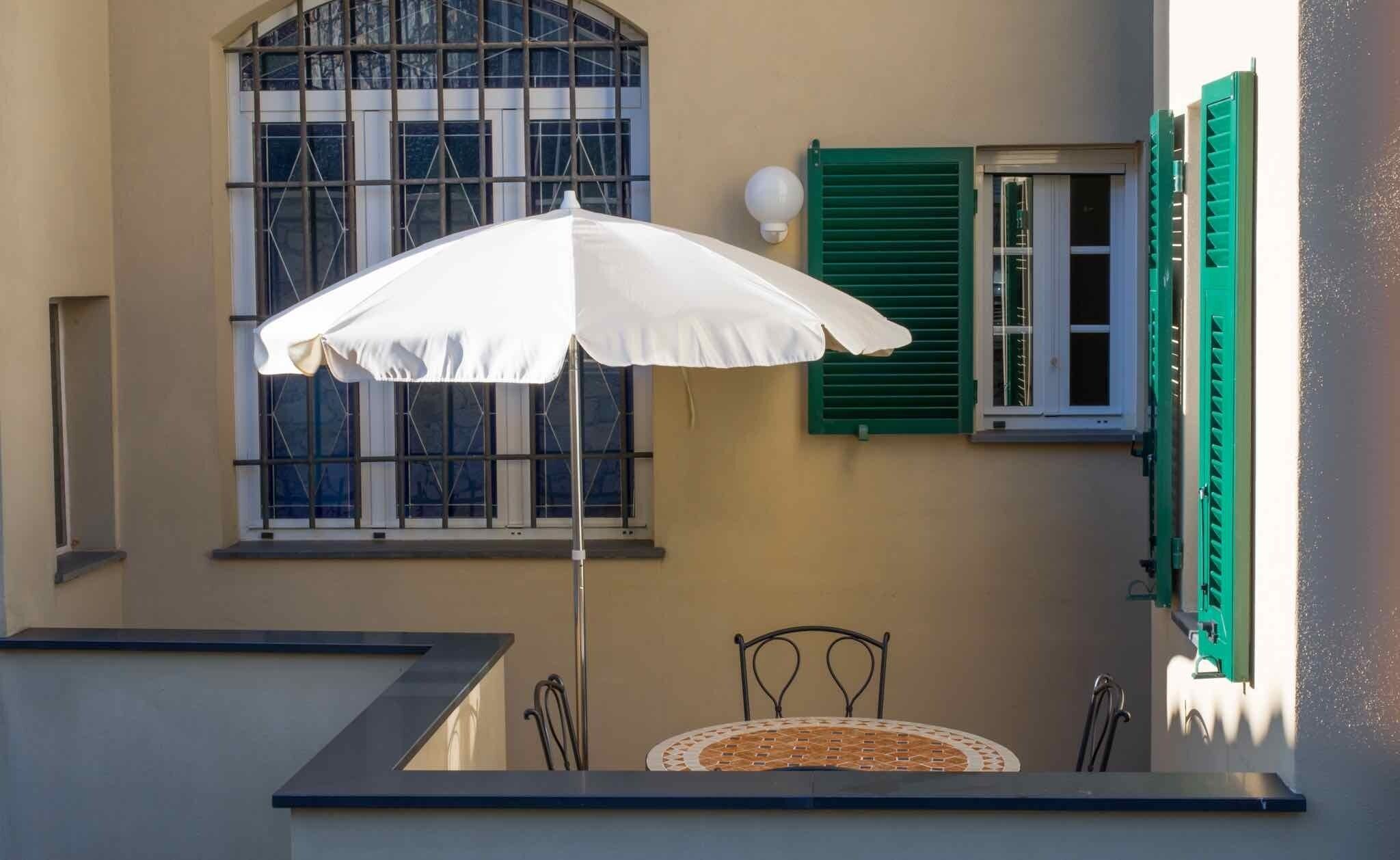 A balcony with a table and chairs, a white umbrella, and a window with green shutters.