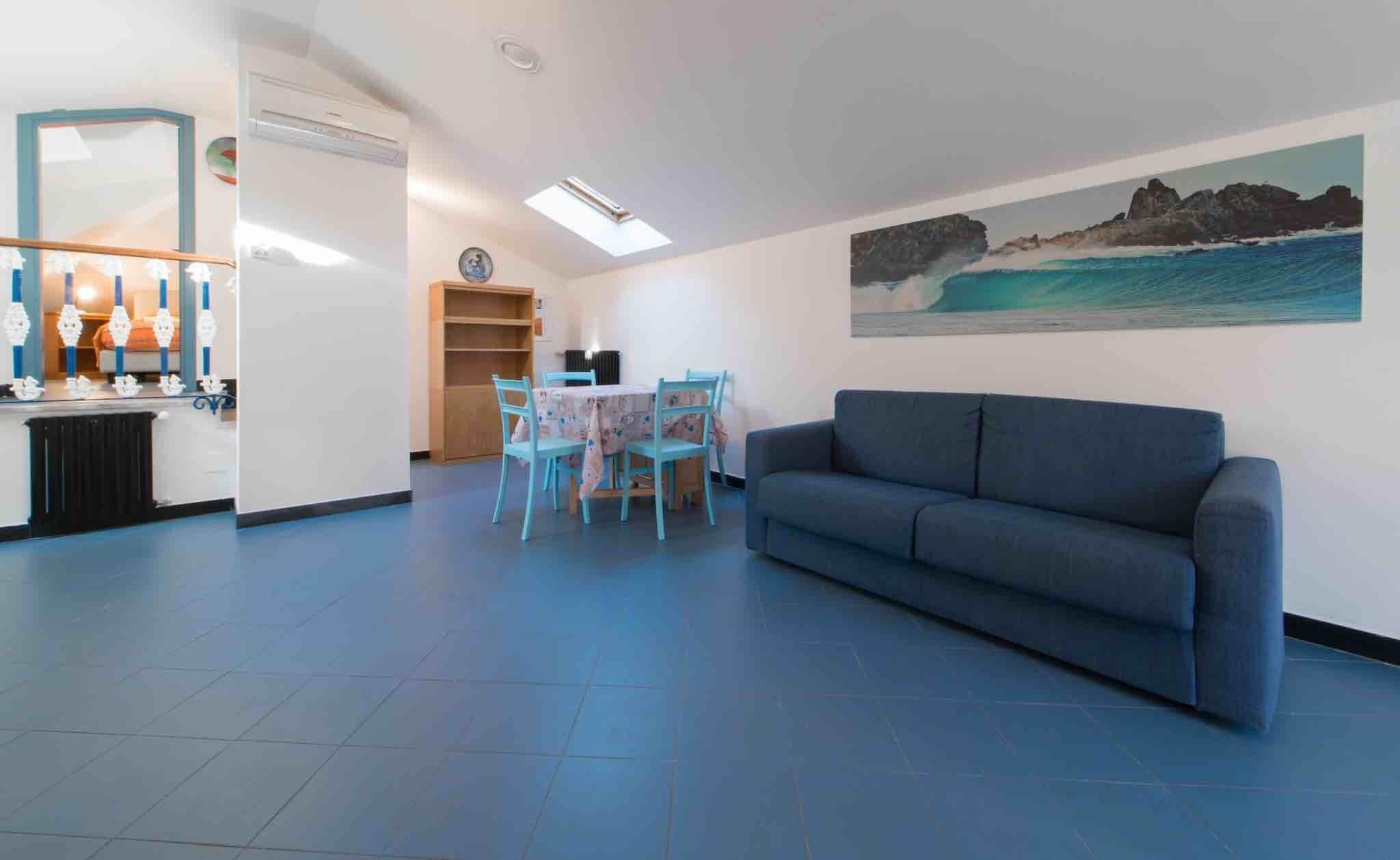 A living room with a sofa, a dining table with chairs, a kitchen area with a fridge, and a painting of a coastal scene.